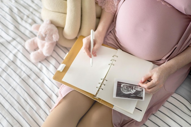 Vista superior carinhosa futura mãe escrevendo diário com foto de ultrassom de raio-x sentada em uma cama confortável
