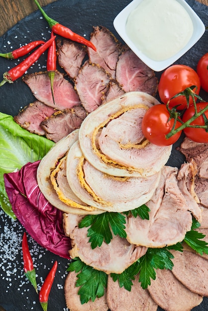 Vista superior de carnes frías, carne asada, jamón al horno, rollito de pollo y lengua de ternera, servido sobre una tabla de piedra