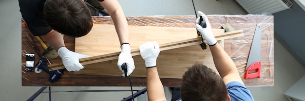 Vista superior de carpinteros masculinos poniendo bloque de madera en tornillo. Uniones que utilizan herramientas especiales de carpintería para el montaje de muebles. Hombres trabajando con tablones de madera. Concepto de taller de carpintería