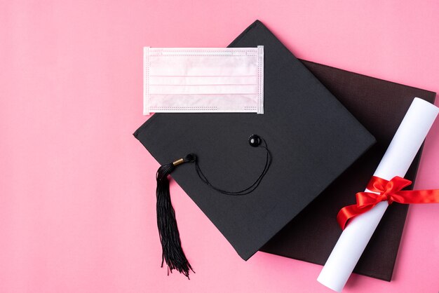 Foto vista superior del casquillo académico cuadrado de graduación con diploma de grado y máscara aislada sobre fondo de tabla rosa.