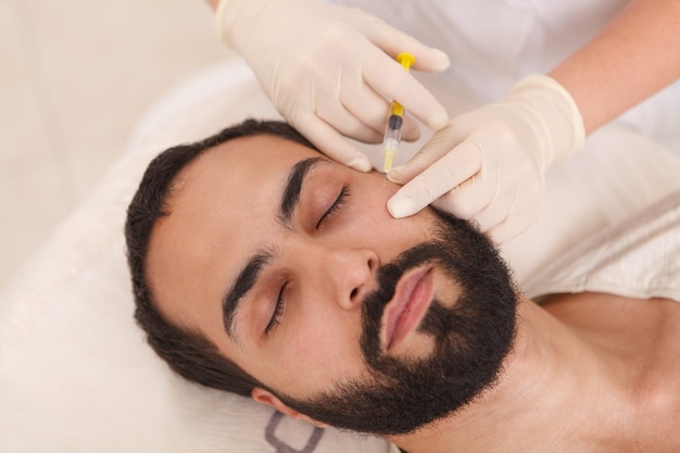 Vista superior de cerca de un hombre barbudo recibiendo inyecciones de relleno anti-envejecimiento en la clínica de belleza