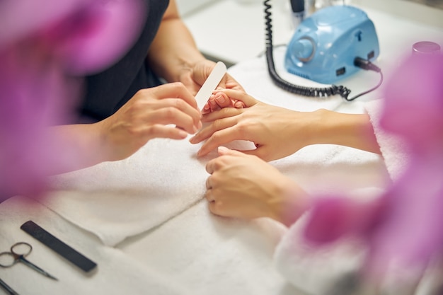 Foto vista superior de cerca de manos femeninas mientras esteticista está puliendo las uñas rodeadas de orquídeas púrpuras