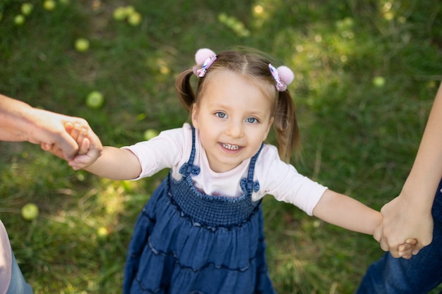 Vista superior de cerca de risa adorable niña de 3 años en vestido de jeans, divirtiéndose y disfrutando de su paseo al aire libre en el parque, tomados de la mano de su madre y su abuela. Paseo familiar en el jardín.