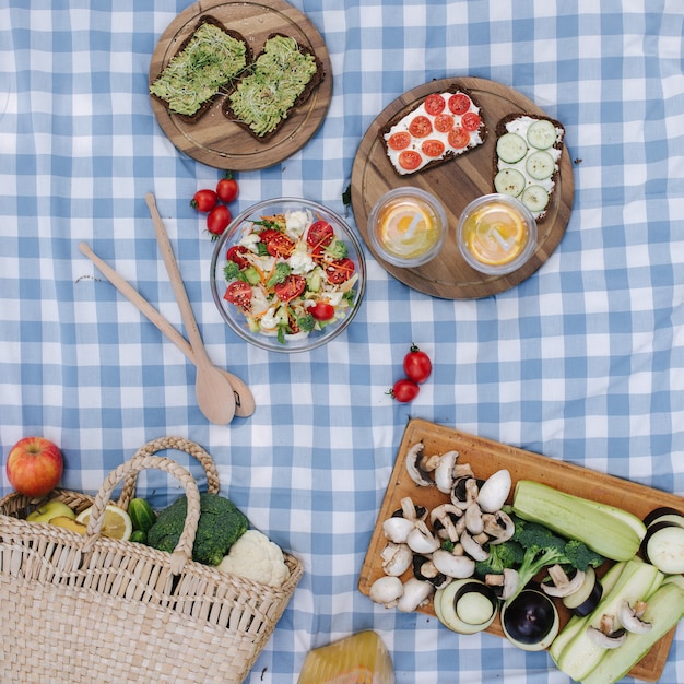 Vista superior de la cesta de picnic con sándwiches veganos saludables en una manta a cuadros azul en el parque