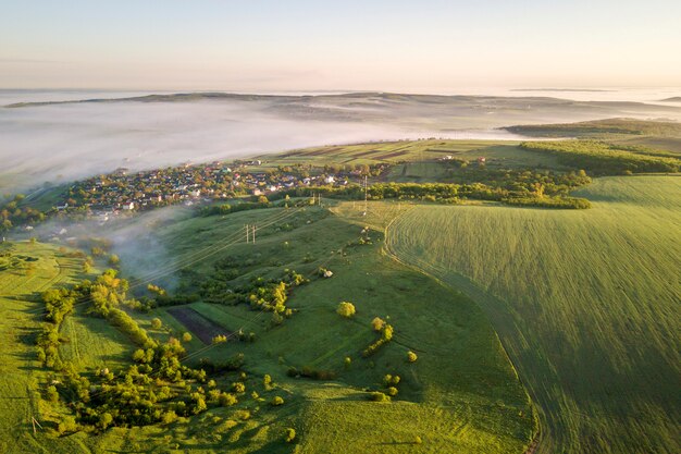 Vista superior de la colina cubierta de hierba verde de niebla, tejados de la casa del pueblo en valle entre árboles verdes en fondo del cielo azul. Panorama de paisaje brumoso de primavera al amanecer.