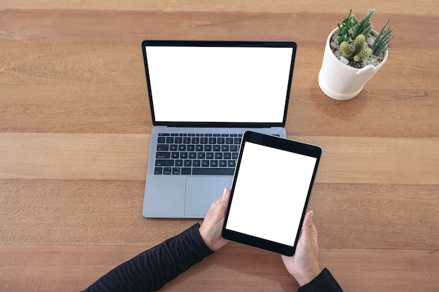 Vista superior com as mãos segurando um tablet preto e um laptop com uma tela em branco na mesa de madeira no escritório