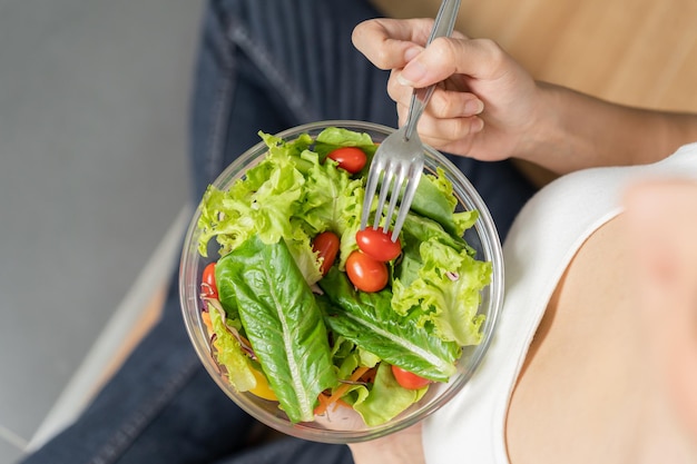 Vista superior comida feliz comiendo un desayuno saludable en la mañana mujer joven asiática con ensalada fresca tomate verde lechuga cos en un tazón sentado en el piso en la cocina Dieta concepto de comida vegetariana