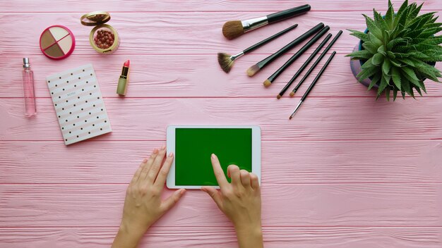 Vista superior de la composición creativa con cosméticos, herramientas de maquillaje, accesorios y manos de mujer usando tableta en superficie de color. Concepto de belleza y moda.