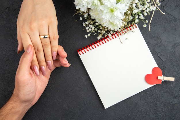 Vista superior concepto de propuesta de matrimonio mano masculina sosteniendo la mano femenina con el cuaderno de flores de anillo sobre fondo oscuro