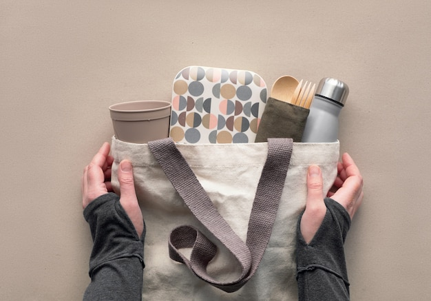 Vista superior criativa, kit de almoço embalado sem desperdício em saco de lona. Mãos segurando um saco com lancheira para viagem, pacote com talheres de bambu, caixa reutilizável e xícara de café para viagem. Layout plano em papel ofício.