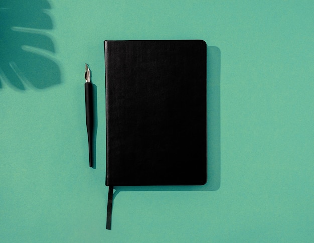 Vista superior del cuaderno y bolígrafos negros de estilo minimalista sobre fondo verde, superposición de sombra de palmera