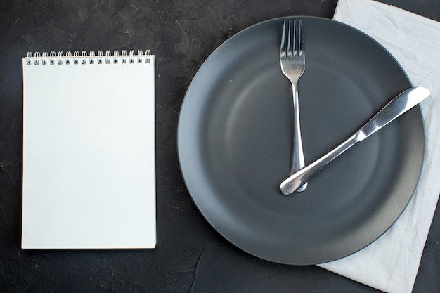 Vista superior del cuaderno y cubiertos en un plato negro sobre una toalla blanca sobre fondo de color oscuro con espacio libre