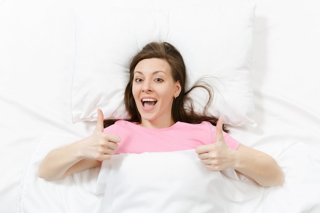 Vista superior da cabeça de uma jovem morena feliz deitada na cama com lençol branco, travesseiro e cobertor