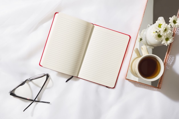 Vista superior da cena da manhã em fundo de tecido voile com um caderno vermelho, óculos, caneca de chá e um vaso de flores brancas em uma bandeja de latão espelhada, com espaço para texto
