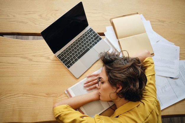 Vista superior da jovem cansada adormece na mesa com laptop e documentos sob a cabeça no local de trabalho