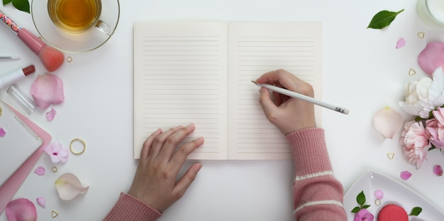 Vista superior da jovem escrevendo no caderno em branco no espaço de trabalho feminino rosa doce com maquiagem