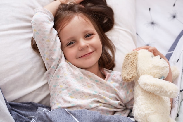 Vista superior da linda garota acordando com seu animal de brinquedo deitado no travesseiro sob o cobertor, olhando sorrindo diretamente para a câmera