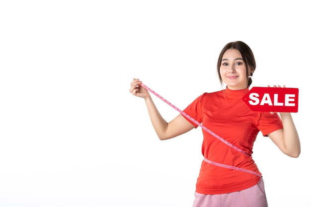 Foto vista superior da linda garota sorridente satisfeita em redorange, blusa segurando um cartão do banco, medindo-se com medidor, segurando a placa de venda no fundo branco