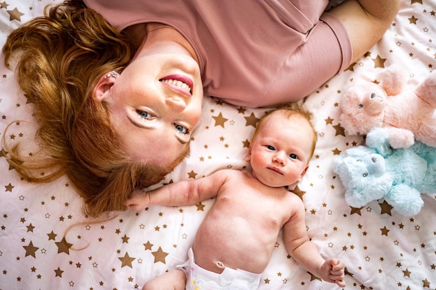 Vista superior da mãe e da fralda engraçada do bebê recém-nascido deitados juntos em uma aconchegante cama de algodão se divertindo