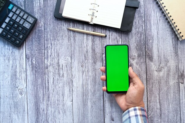 Vista superior da mão de um homem segurando um telefone inteligente com tela vazia na mesa do escritório