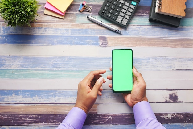 Vista superior da mão de um homem segurando um telefone inteligente com tela vazia na mesa do escritório