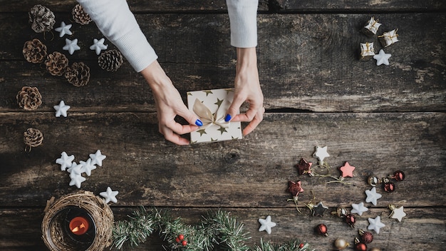 Vista superior da mão feminina amarrando o laço dourado em cima da caixa de presente de feriado em uma mesa de madeira rústica com enfeites de natal, decoração e velas colocadas por toda parte.