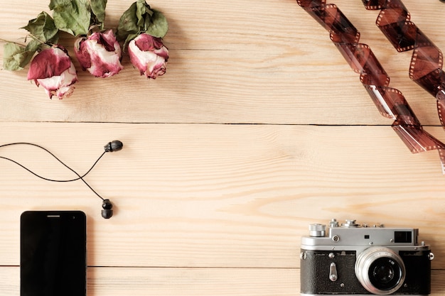 Vista superior da mesa de madeira com smartphone, fones de ouvido, câmera vintage, filme e rosas secas com folhas
