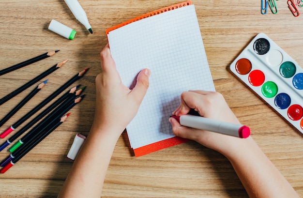 Foto vista superior da mesa de uma criança em idade escolar com material de escritório, lápis, canetas hidrográficas, tesouras, tintas e a escrita de uma criança em um caderno