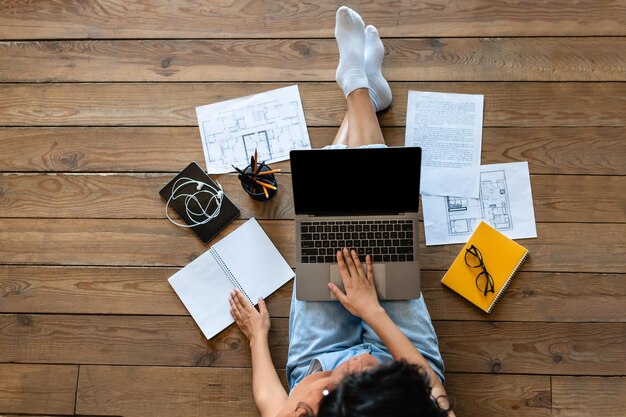 Vista superior da mulher com materiais de trabalho ou estudo usando laptop com tela vazia enquanto está sentado no chão