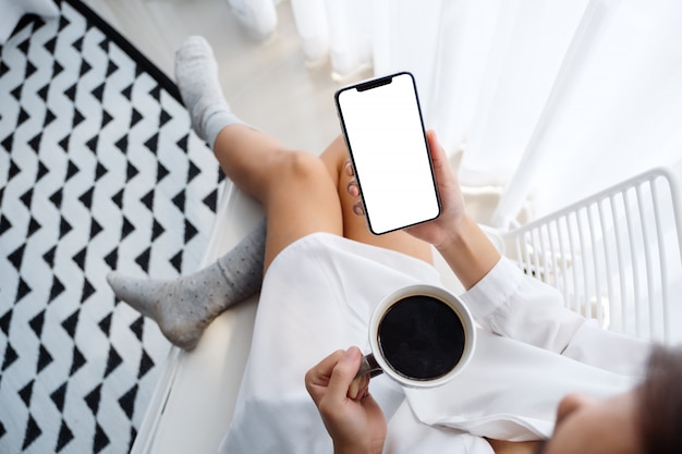 Vista superior da mulher segurando o telefone móvel com tela branca em branco enquanto está sentado e tomando café no quarto