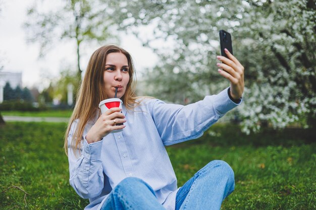 Vista superior da mulher sentada no jardim do parque na grama verde com livro de tablet de fones de ouvido de smartphone e café na mão