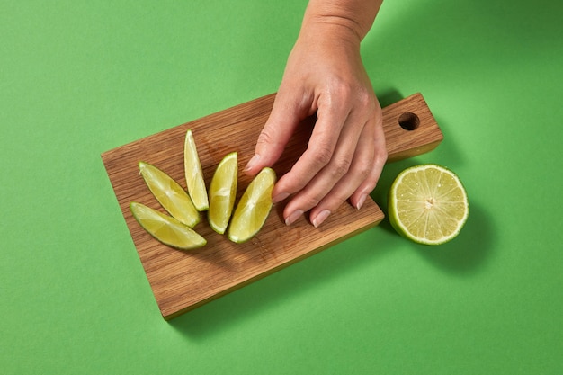 Vista superior da parede cítrica. fatias de limão em uma placa sobre uma mesa verde. uma mão feminina coloca uma fatia de limão verde fresco maduro em uma tábua de madeira com espaço de cópia.