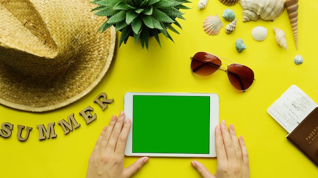 Vista superior das mãos de mulher segurando um tablet digital com tela verde, fundo de acessórios de verão