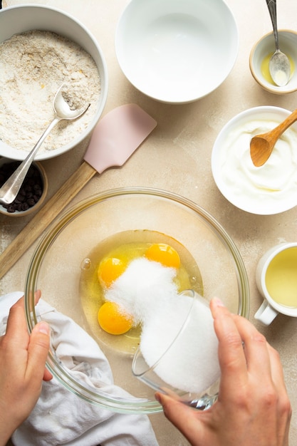 Vista superior das mãos femininas derramando açúcar sobre os ovos na tigela de vidro. Receita passo a passo.