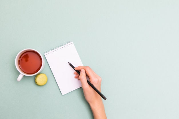 Vista superior das mãos femininas, escrevendo em um bloco de notas em branco na recepção. na mesa azul, havia uma xícara de chá e biscoito.