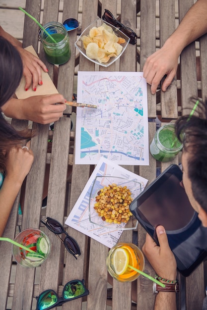 Vista superior de amigos jovens turistas que procuram mapas em uma viagem de férias sobre uma mesa de madeira com lanches e bebidas saudáveis. Férias e conceito de turismo.