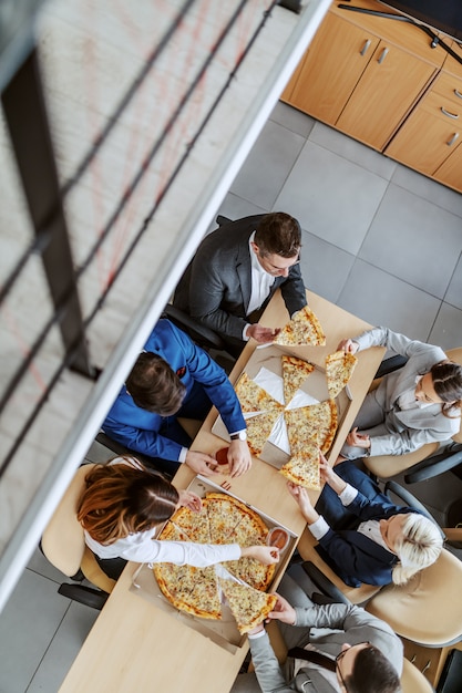 Vista superior de colegas famintos, sentados à mesa e comendo pizza no almoço. Interior da empresa corporativa.