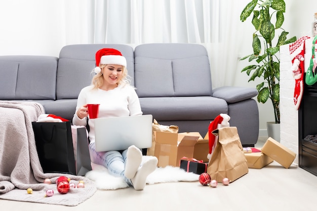 Vista superior de compras online de natal. compradora com laptop, copie o espaço na tela. mulher toma café, compra presentes, prepara-se para a véspera de natal, sentada entre caixas e pacotes de presentes. promoções de férias de inverno