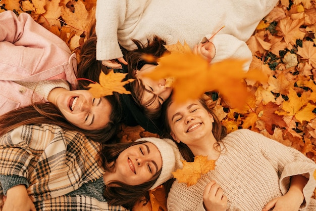 Vista superior de jovens amigas sorridentes de cabelos louros e cabelos escuros, dois deles com chapéus na cabeça, vestidos com jaquetas brancas, cinza, rosa e bege, deitadas em folhas de outono laranja no parque