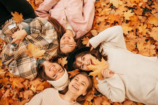 Vista superior de jovens amigas sorridentes de cabelos louros e cabelos escuros, dois deles com chapéus na cabeça, vestidos com jaquetas brancas, cinza, rosa e bege, deitadas em folhas de outono laranja no parque