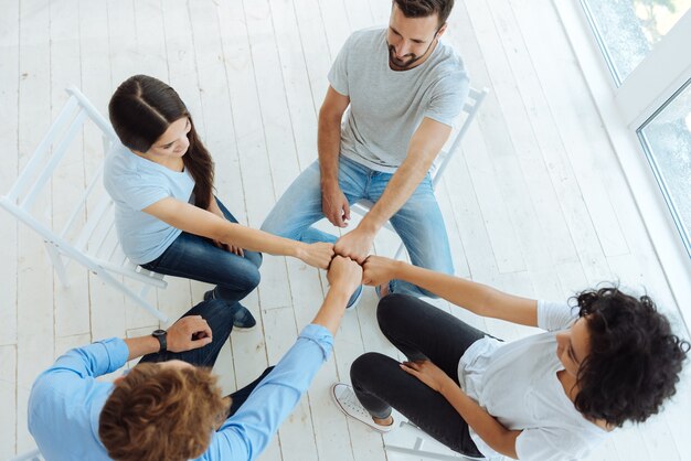 Vista superior de jovens positivamente maravilhados, sentados em círculo e sorrindo enquanto mantêm os punhos fechados