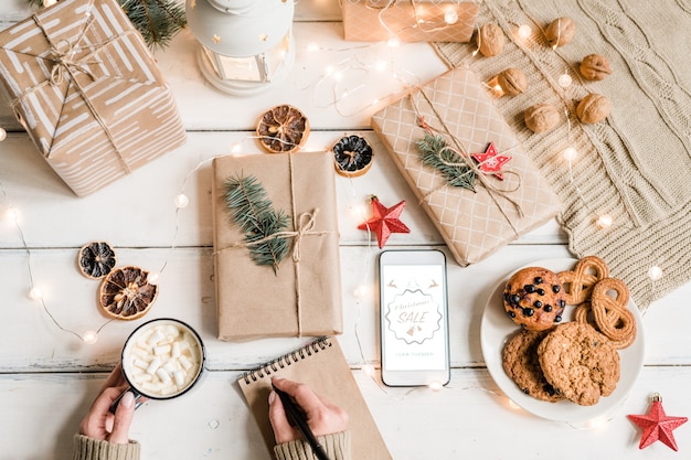 Vista superior de mãos femininas com caneta sobre o caderno segurando uma caneca com bebida quente entre presentes embalados, biscoitos e decorações de Natal