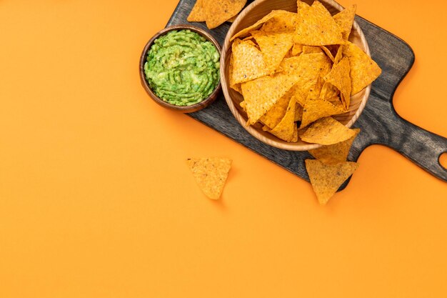 Vista superior de nachos mexicanos crocantes servidos com guacamole na tábua de madeira em fundo laranja
