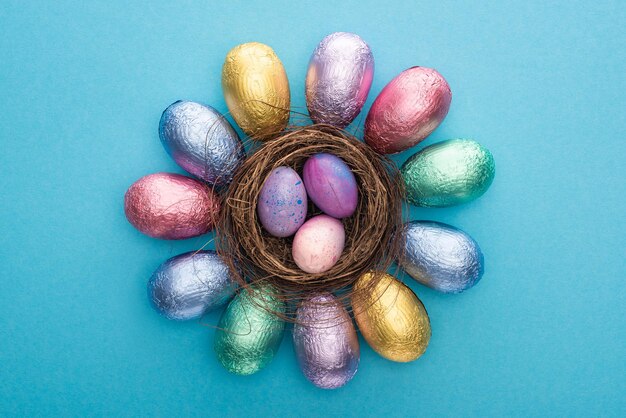 Foto vista superior de ovos de páscoa de chocolate em folha colorida ao redor do ninho com ovos de codorniz em fundo azul