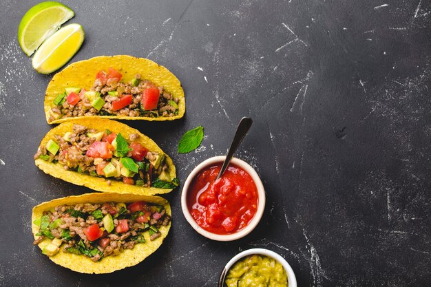 Vista superior de tacos mexicanos com carne, vegetais em tortilla de milho amarelo servidos com molho de salsa e guacamole em fundo de pedra rústica preta