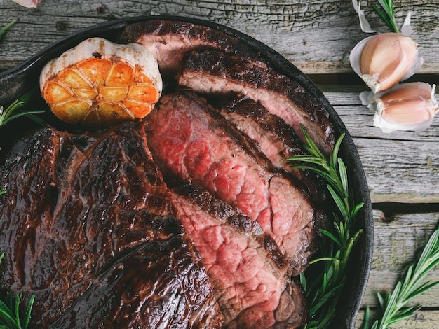 Vista superior de um bife meio raro com alho assado e alecrim em uma panela preta sobre mesa rústica