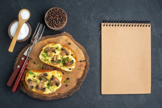 Vista superior de um delicioso lanche com talheres de cogumelos no quadro de madeira e caderno de especiarias em fundo preto