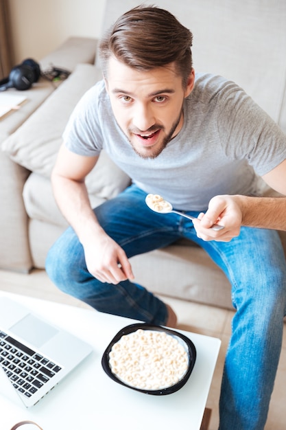 Vista superior de um homem barbudo feliz sentado no sofá comendo cereais em casa