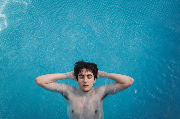 Vista superior de um jovem com os olhos fechados, flutuando em uma piscina com as mãos na nuca. Aproveitando as férias enquanto se bronzeava em um hotel. Conceito de férias.