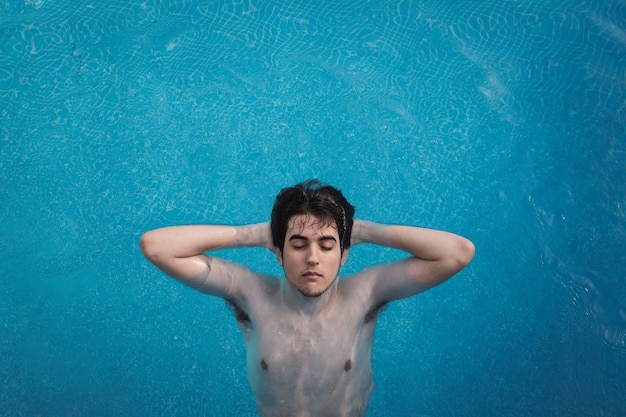 Vista superior de um jovem com os olhos fechados, flutuando em uma piscina com as mãos na nuca. Aproveitando as férias enquanto se bronzeava em um hotel. Conceito de férias.